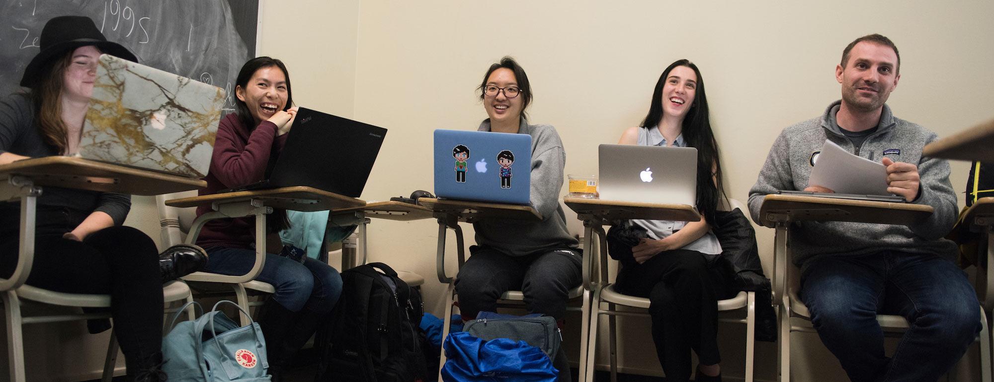 Students sit at desks smiling behind their laptops