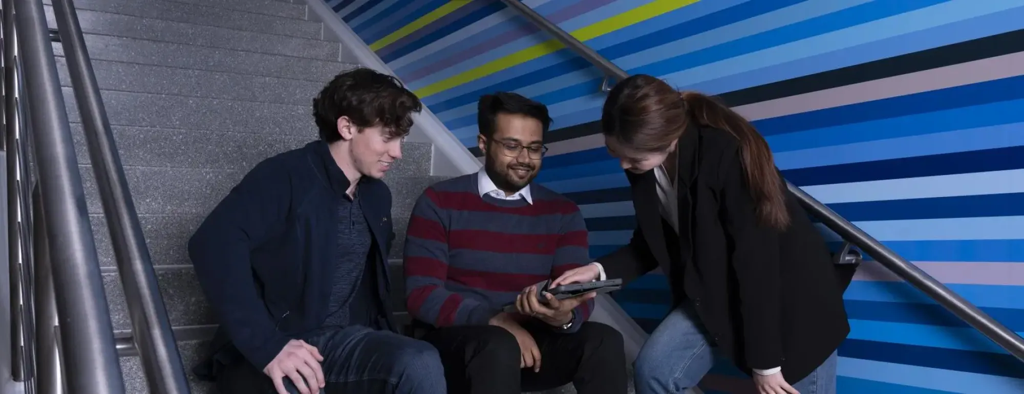 three students sitting on steps talking and looking at a device