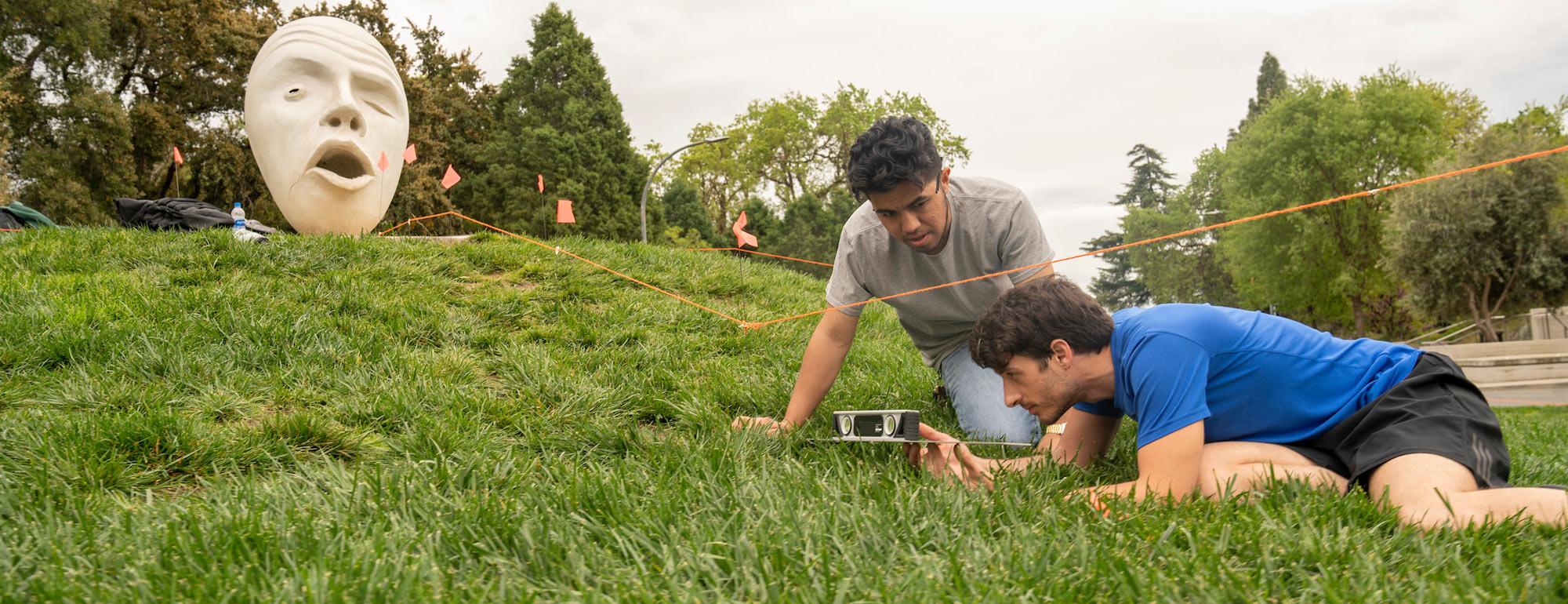 Two male students survey a landscape site with one of the UC Davis eggheads in the background