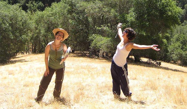 Two women stand on a grassy knoll with trees behind them