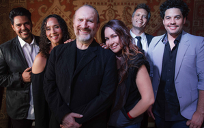 Colin Hay wearing black and sporting a goatee, stading in front of the members of his band.