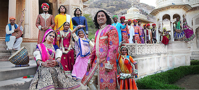 The band members standing in front of an Indian builing, wearing brightly colored costumes.