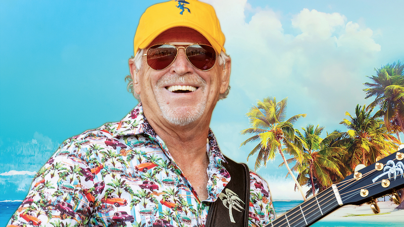 Jimmy Buffett smiling, holding his guitar, wearing sunglasses and a yellow baseball cap.