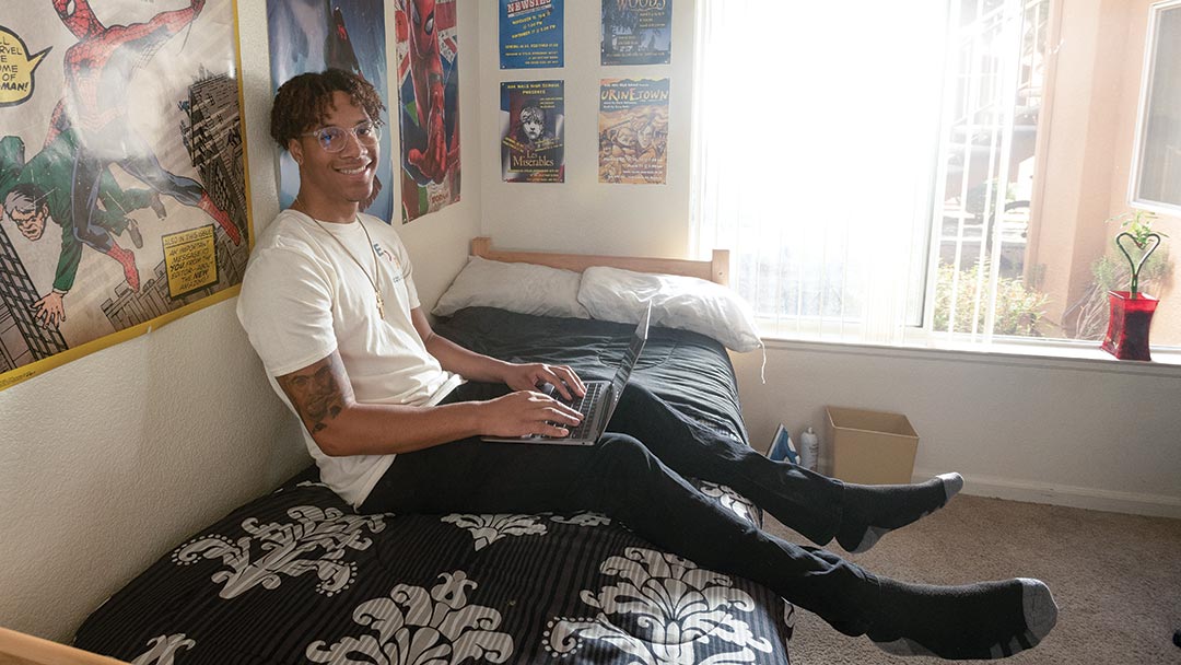 A student sits on his bed with a laptop