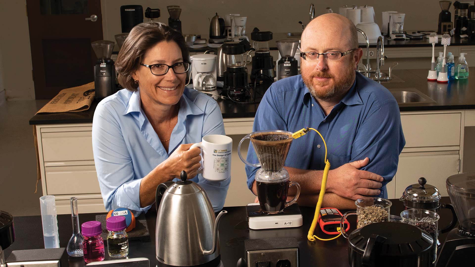 Professors Tonya Kuhl and Bill Ristenpart sit side by side facing us from behind the counter of a lab station. Kulfis is holding a cup of coffee and Ristenpart is sitting with crossed arms on the table, while an instrument is attached to a coffee filter with a yellow cord as the coffee drips into a clear glass cup. Various coffee makers and lab instruments are in front of and behind them.