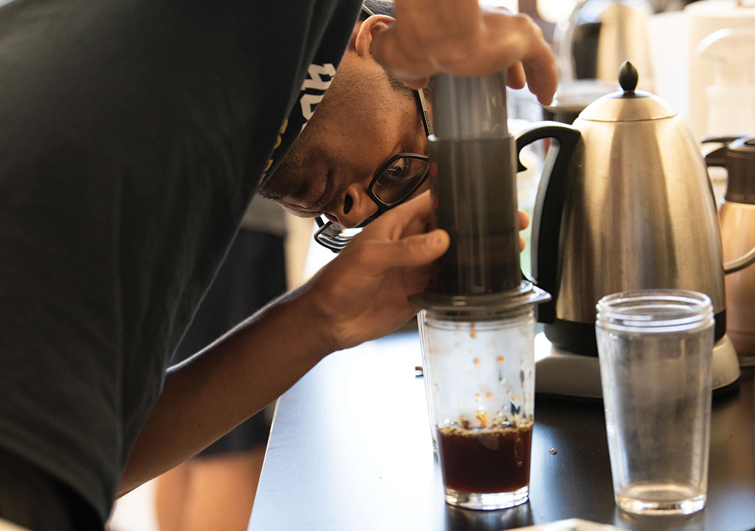 A coffee class student bends down to look carefully at a glass sitting under a filter with coffee in it as it drips into the glass.