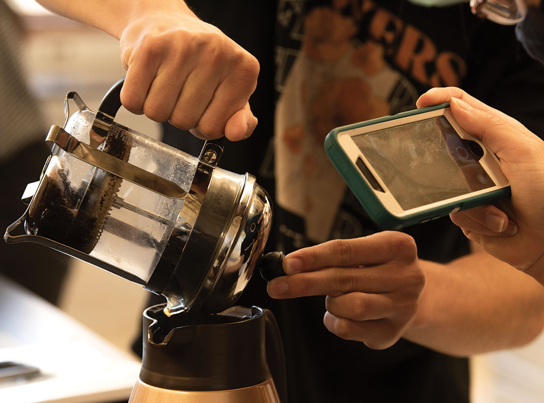 Closeup photo of a student's hands as coffee is being poured from a French press into a carafe. Another student's hands are shown holding a smartphone and taking video of the pour.