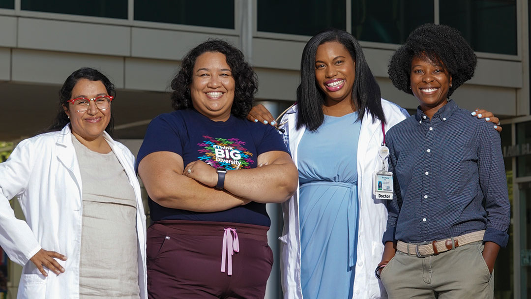 A group of four women stand together in front of a medical office building. Two wear lab coats and one wears scrubs. One has a stethoscope. All are smiling and looking directly at us.