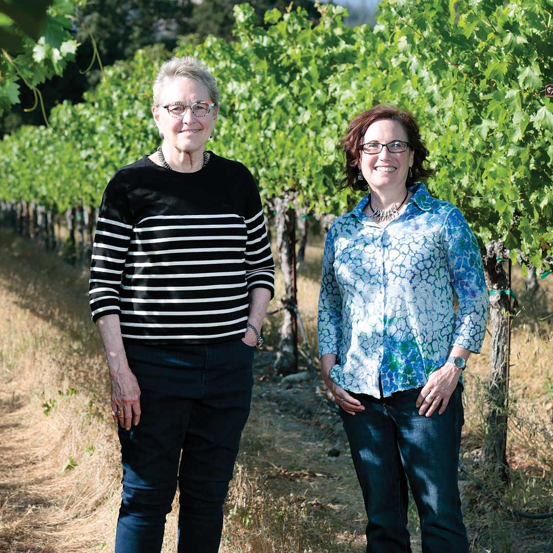 Two women stand next to each other in a sunny vineyard, both of them smiling at looking at us.