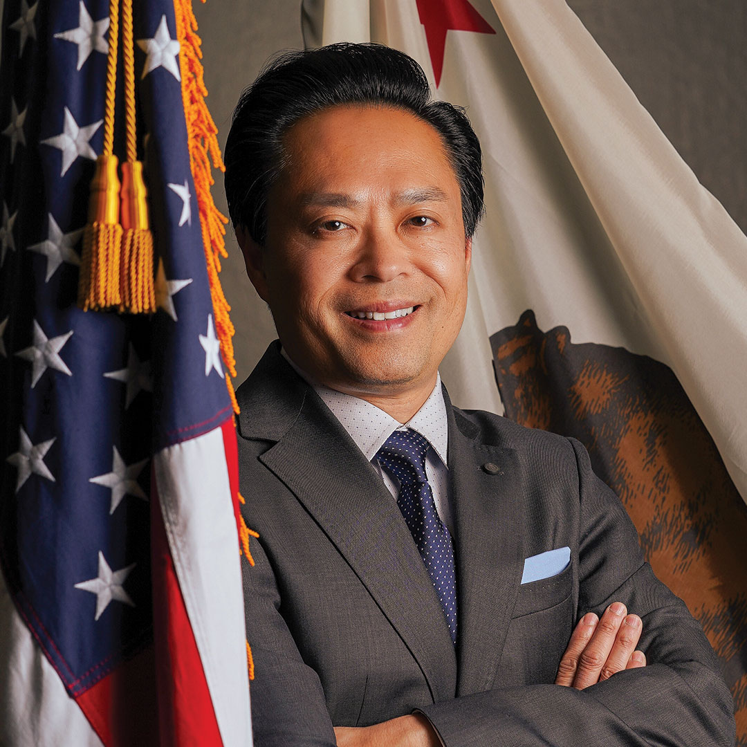 Portrait of a man wearing a gray suit and standing in front of the U.S. and California flags, with his arms crossed and smiling.