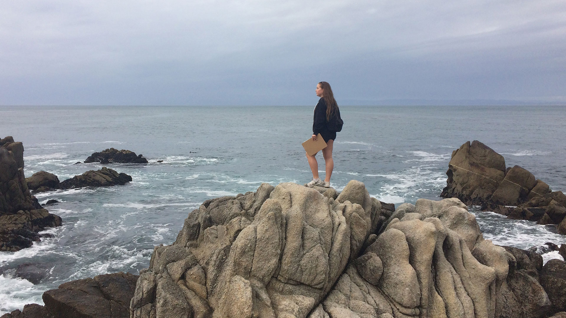 A woman stands on giant shoreline rocks looking out at the ocean and holding a clipboard