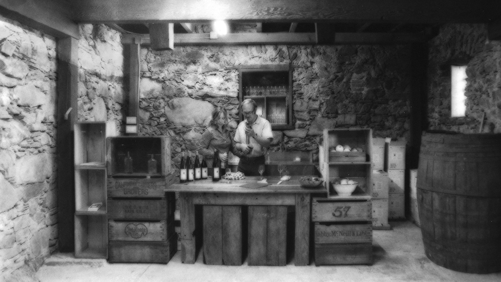 An old black and white photo of a man and woman behind a counter in a room with stone walls and exposed timber beams. A large wine barrel is off to the side.
