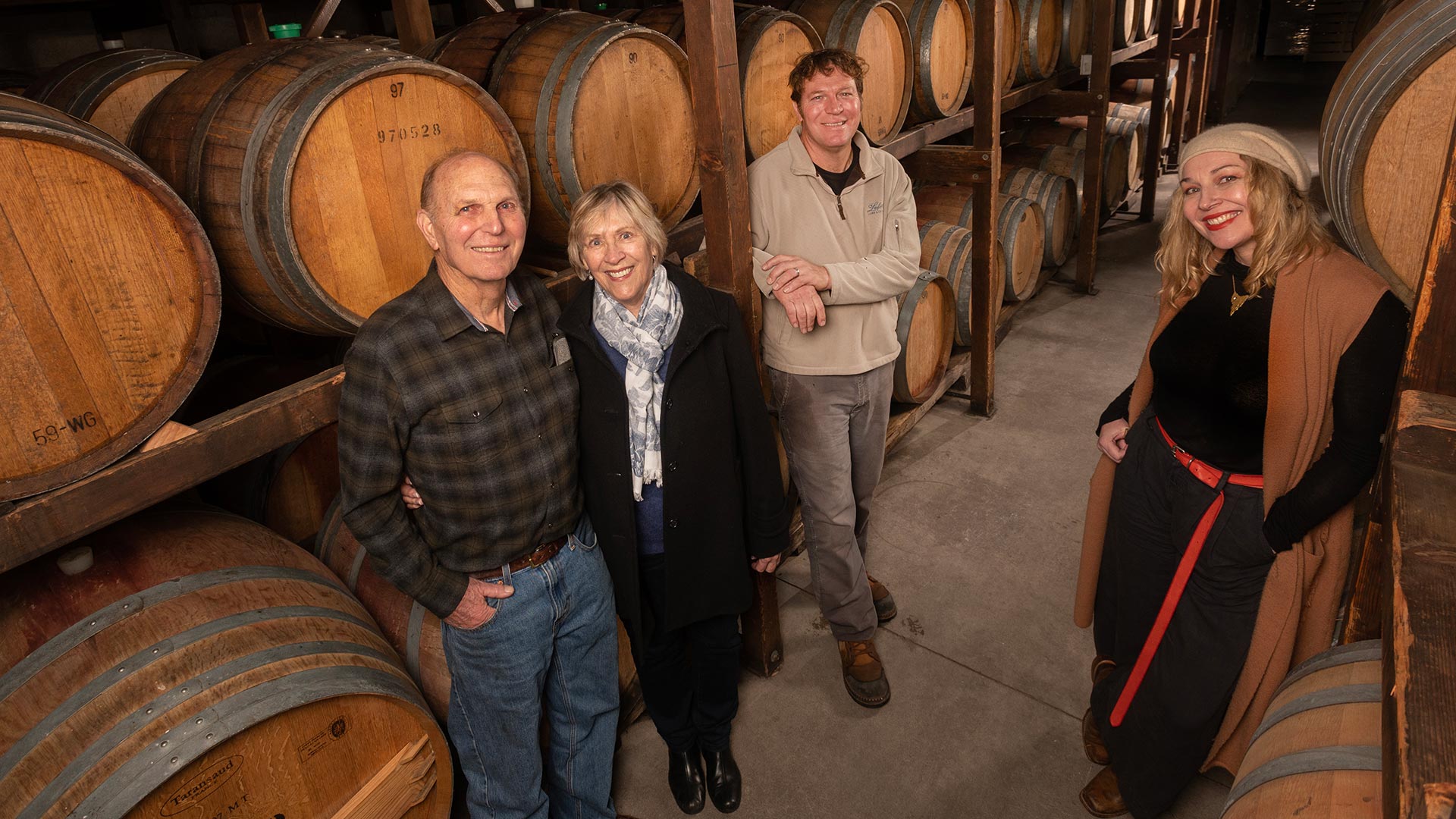 Four people stand in a wine cellar while looking at the camera. Wine barrels are racked in rows on both sides of them.