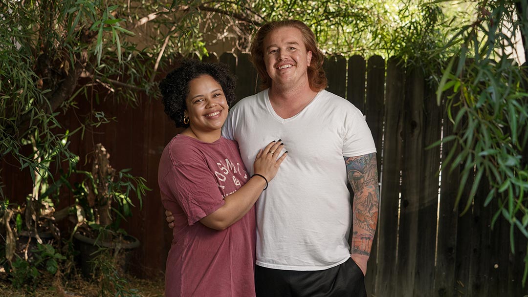 Portrait of a man and a woman standing together under trees and in front of a fence. He wears a white t-shirt and she wears and maroon t-shirt.