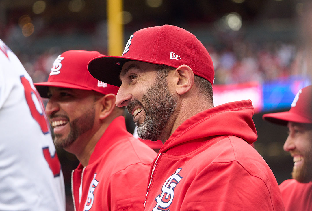 A man in a red cap and jacket smiles looks off camera, smiling. He is in a dugout with other people wearing red.