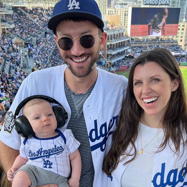 A man, woman and baby pose for a photo. All three are wearing Dodgers jerseys and hats, and are in the stands at a large baseball stadium.