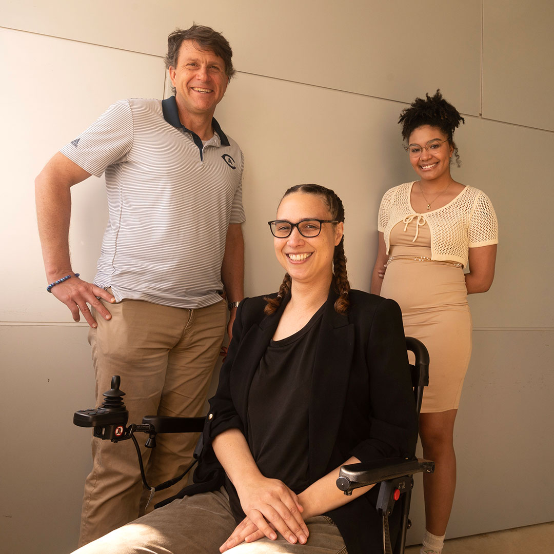 Three people pose for the camera. The person on the left stands with a hand on their hip, while a second sits in a wheelchair and a third stands to the right, hands behind their back.