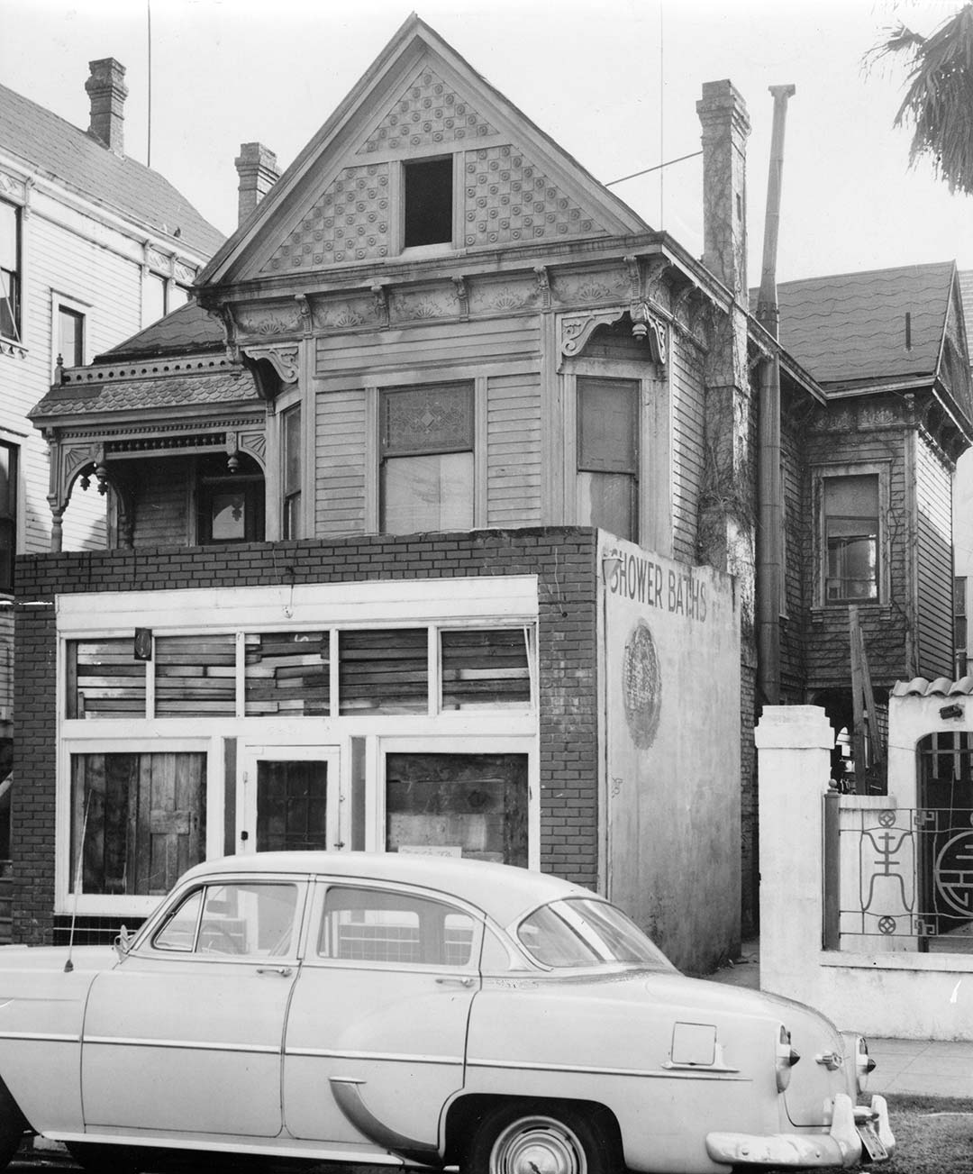 Black and white photo of a three story house. The house has a peaked roof, bay window in the front on the second floor and a closeed-in porch on the ground floor. A sedan is parked on the street in front. The windows are boarded up.