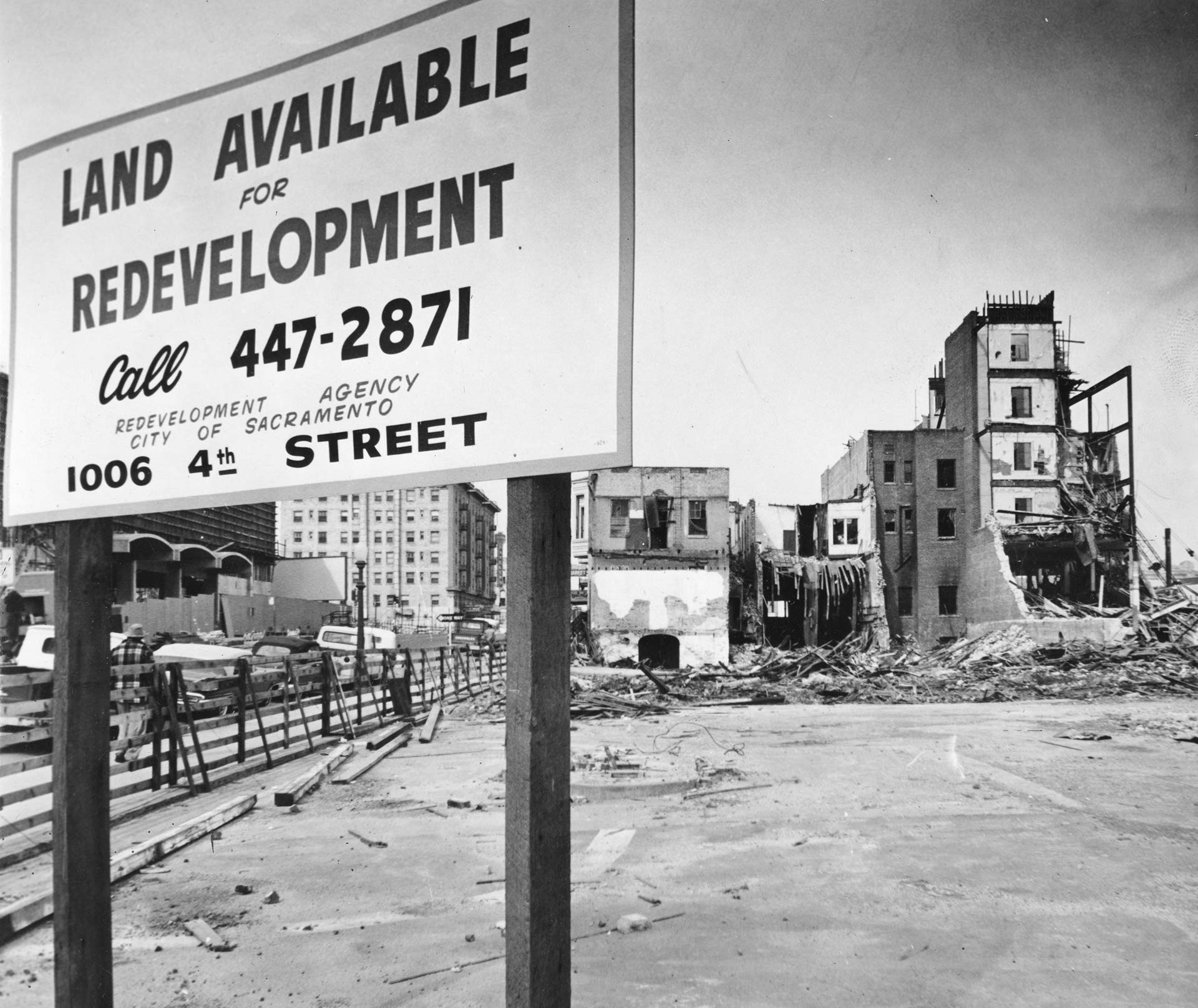 Black and white photo showing a large sign put up in front of a vacant lot. The sign reads "Land Available for Redevelopment" and gives a phone number and address. Buildings toward the back of the lot are undergoing various stages of demolition.