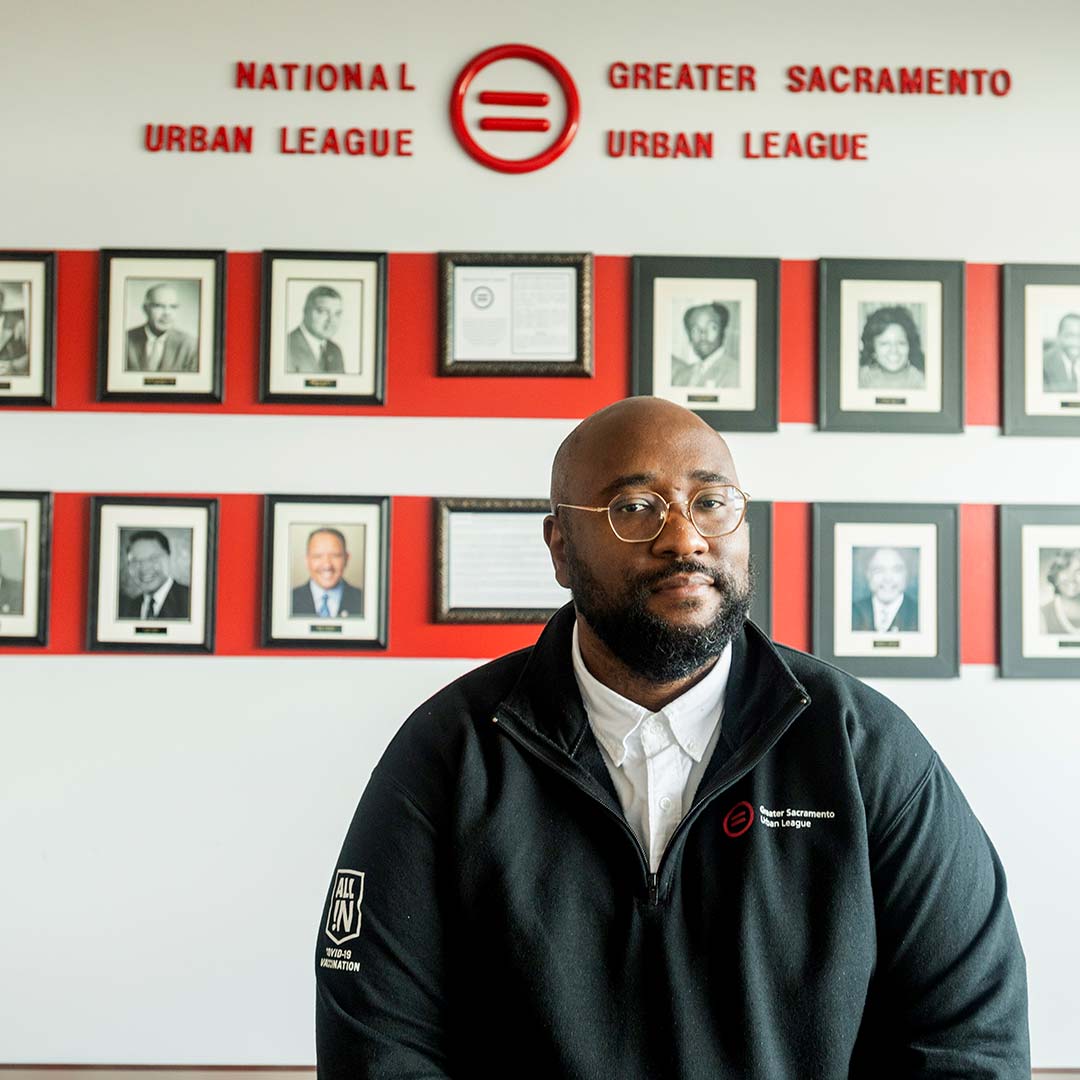 Portrait of a man sitting in front of a wall full of portraits of various people. The sign above him on the wall says National Urban League and Greater Sacramento Urban League. He wears a black jacket, a white button up shirt, has a beard and glasses.